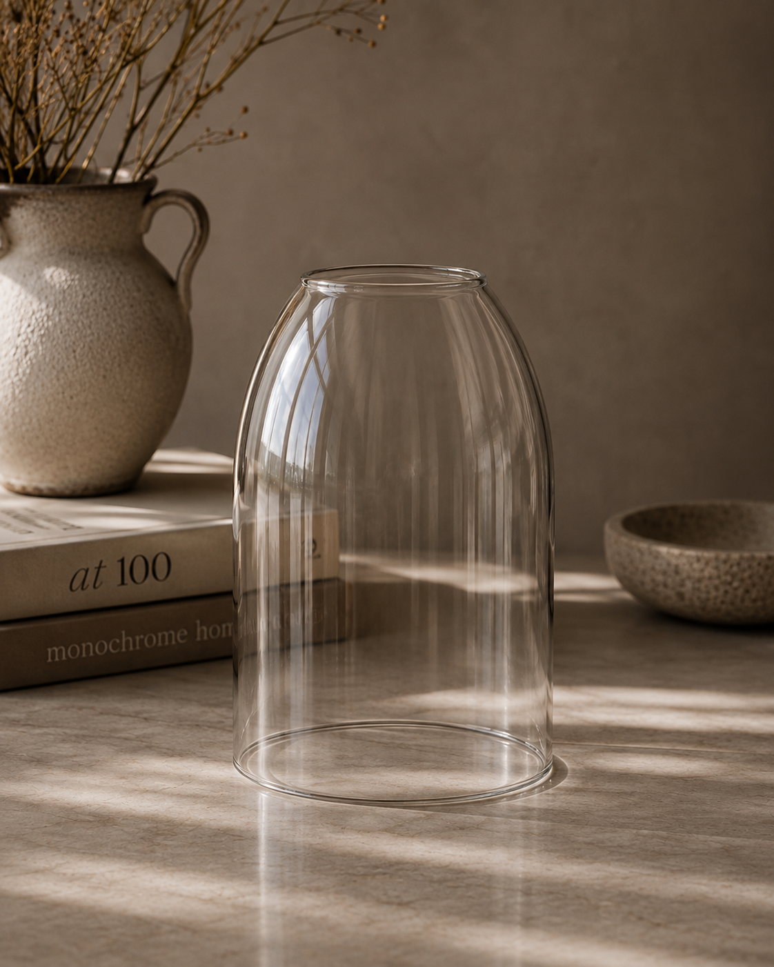 A clear glass bell-shaped vase or planter shown on a tabletop in a styled interior scene with warm sunlight, a ceramic vase with dried branches, stacked books, and a small bowl in the background.