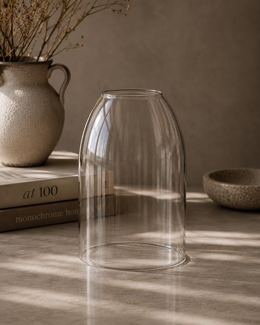 A clear glass bell-shaped vase or planter shown on a tabletop in a styled interior scene with warm sunlight, a ceramic vase with dried branches, stacked books, and a small bowl in the background.