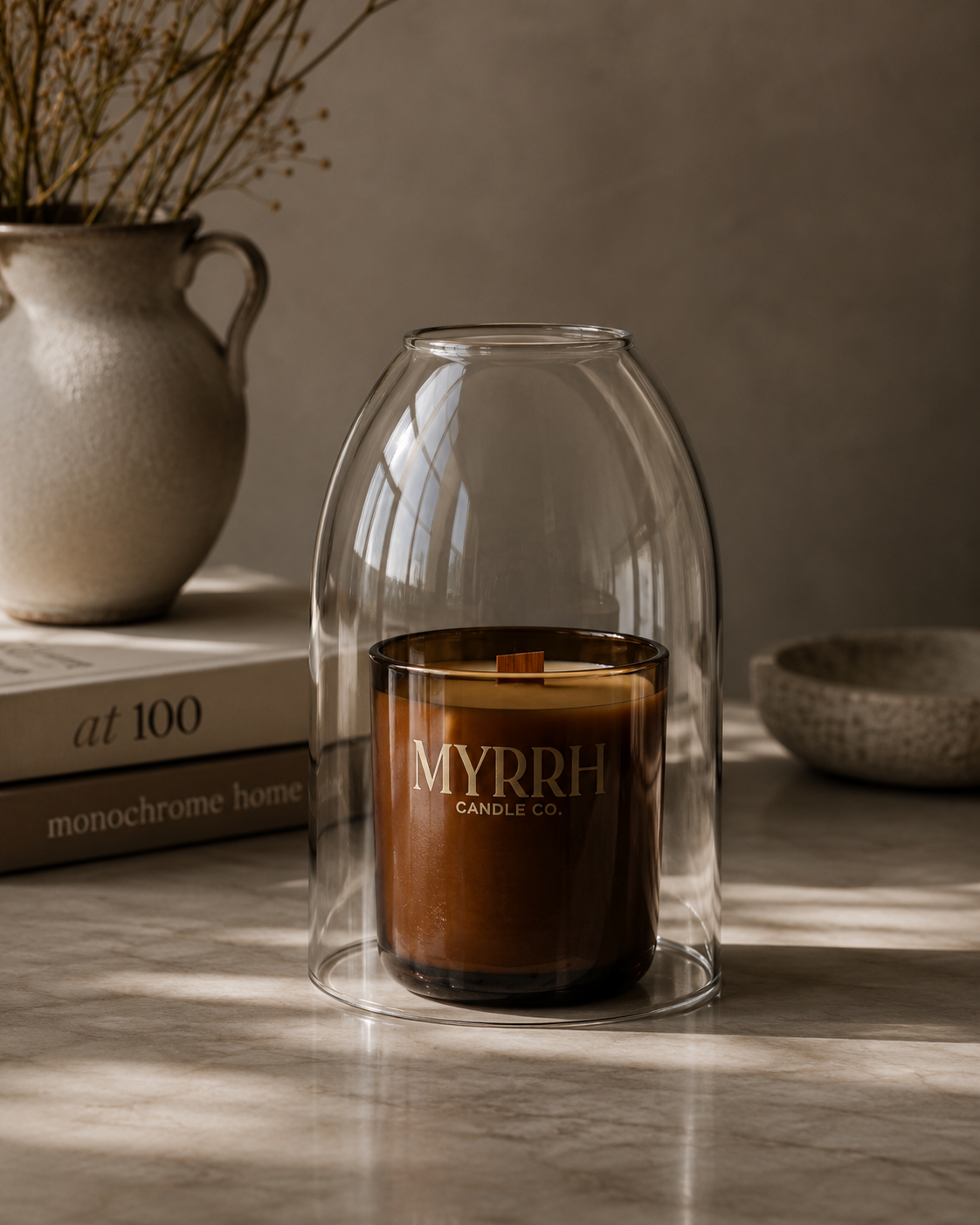 A brown glass jar candle with a single wick sits inside a clear bell-shaped glass cloche. The jar is printed with the brand name 'MYRRH CANDLE CO.